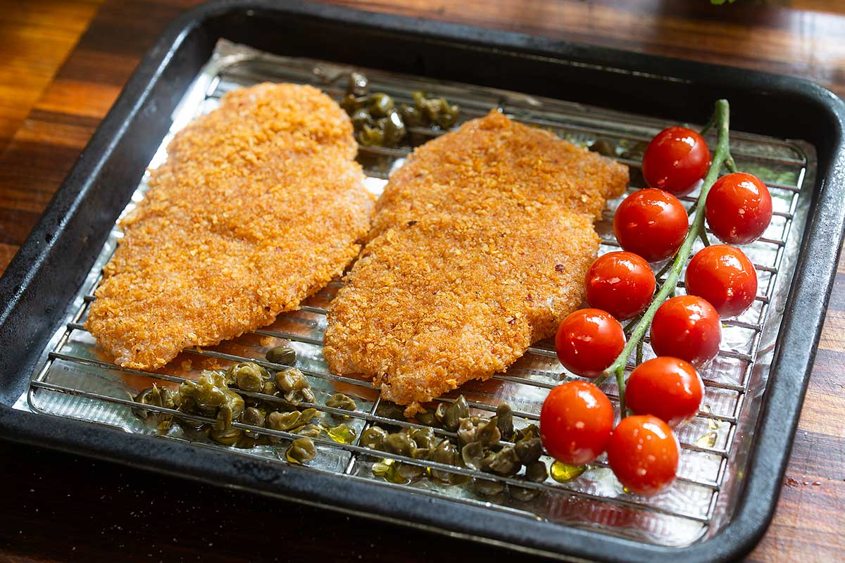 Crumbed cutlets on the oiled wire rack with cherry tomatoes and capers spaced out on the sheet pan]