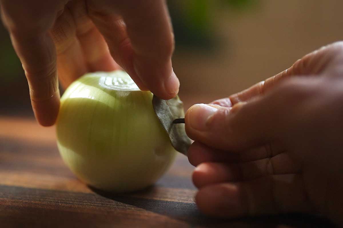 Cloves being pushed into a peeled onion, with the bay leaf pinned on