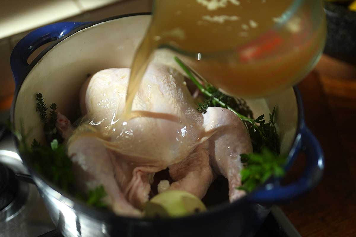 The whole chicken sitting in the pot with the onion, garlic and herbs, stock being poured in